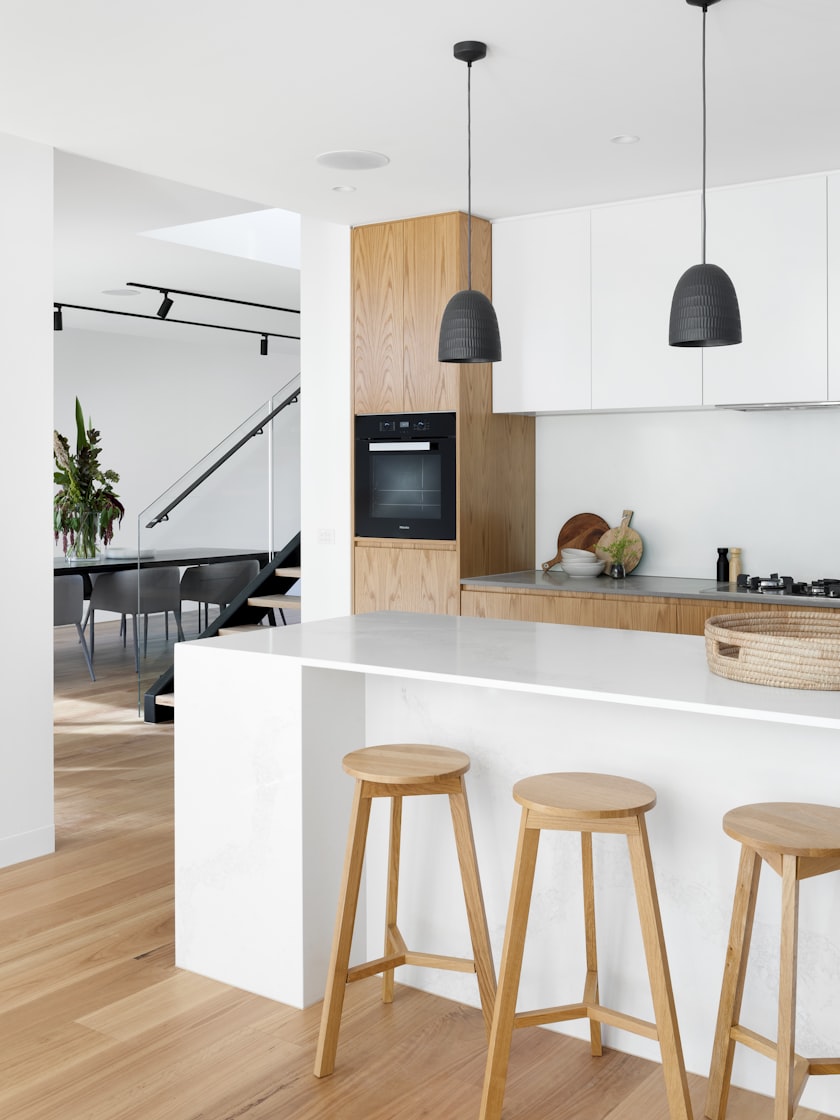 A white kitchen with an island, timber stools and black pendant lights.